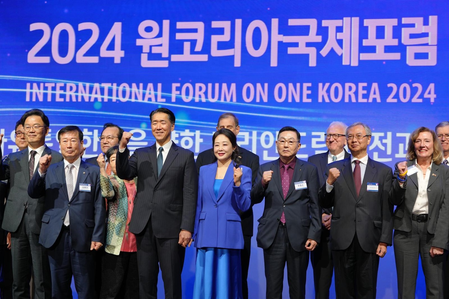 A group of people in formal attire stand together in front of a blue backdrop with the text "2024 International Forum on One Korea" written in English and Korean. Some are raising their fists, embodying the spirit of a Unified Korea.