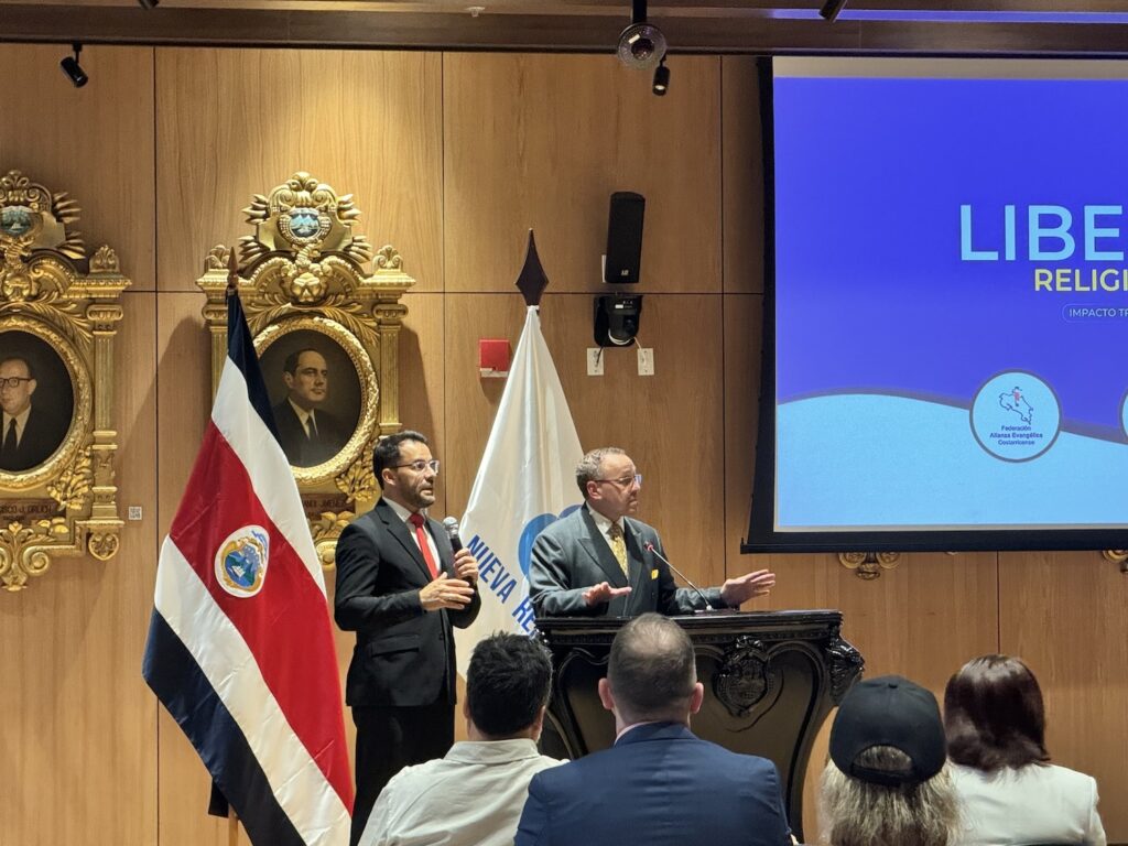 Two men in formal attire give a presentation on religious freedom at a podium in a decorated room with a Costa Rican flag and a projected screen behind them. Audience members are seated in front, attentive to the insights shared by these global leaders.