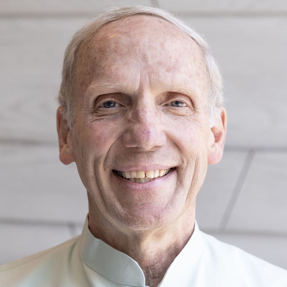 David-Caprara A smiling elderly person with short hair and a light-colored shirt stands in front of a neutral background.