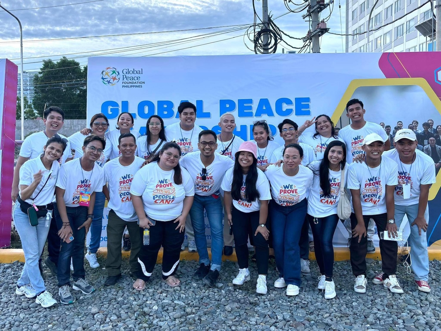 A group of 2024 peacebuilders wearing "We Prove Humanity Can" shirts stands in front of a "Global Peace Foundation" banner, posing for a group photo outdoors as part of the Global Peace Leadership Corps.