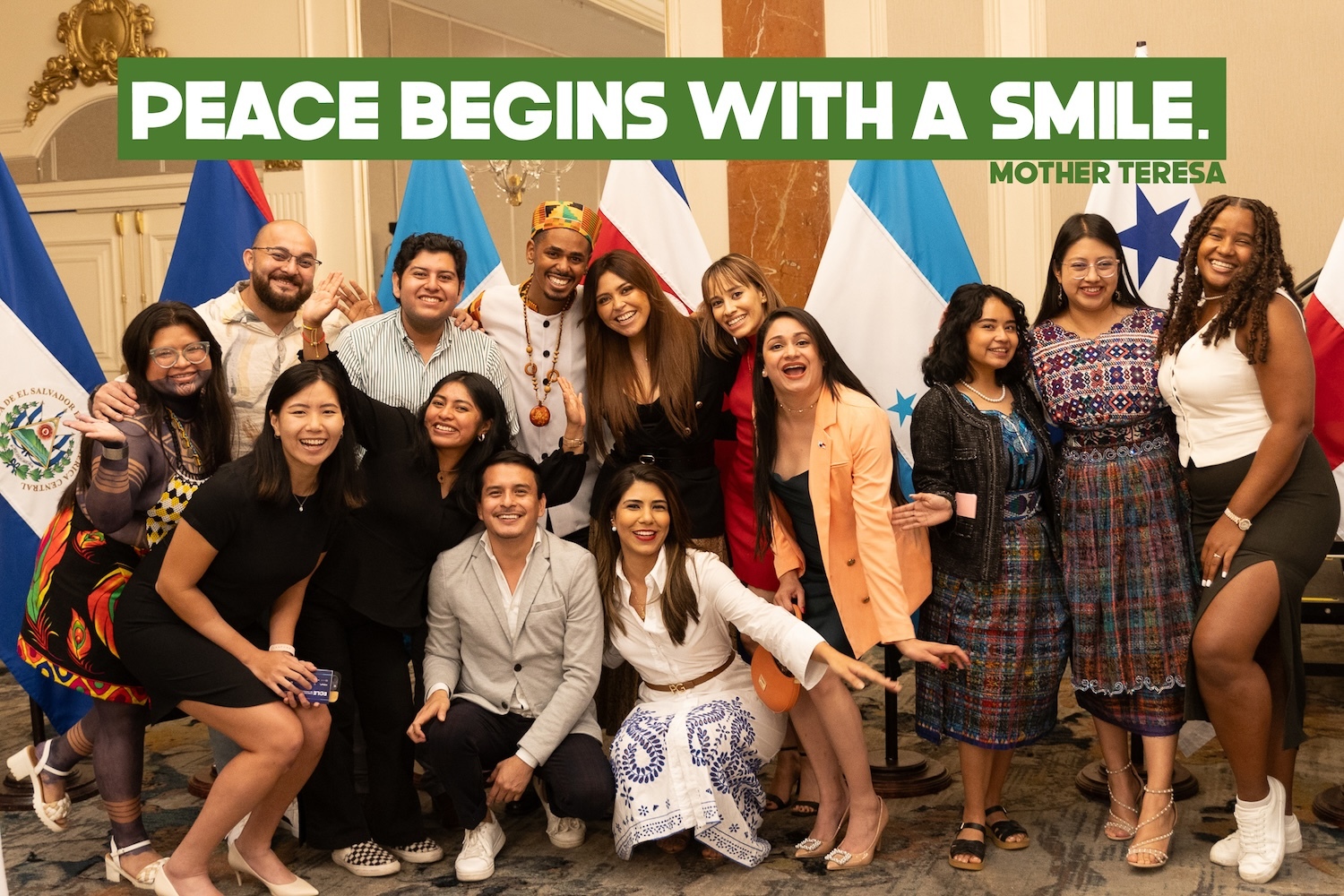 Group of people smiling and posing for a photo in front of various national flags at the Central American Emerging Leaders Summit. Text at the top reads, "Peace begins with a smile." - Mother Teresa.