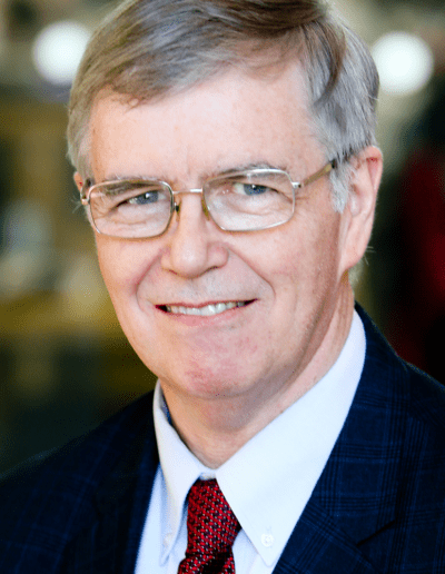 A man with grey hair, glasses, and a plaid blazer smiles softly.