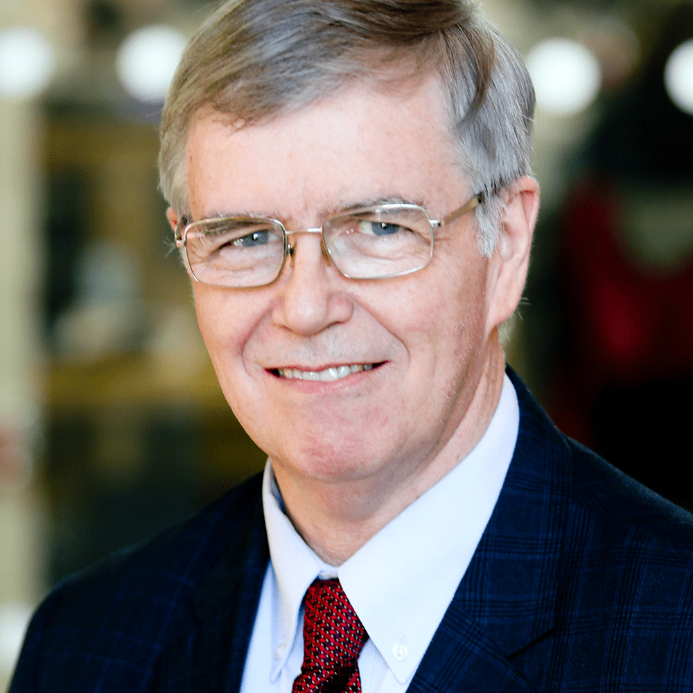 A man with grey hair, glasses, and a plaid blazer smiles softly.