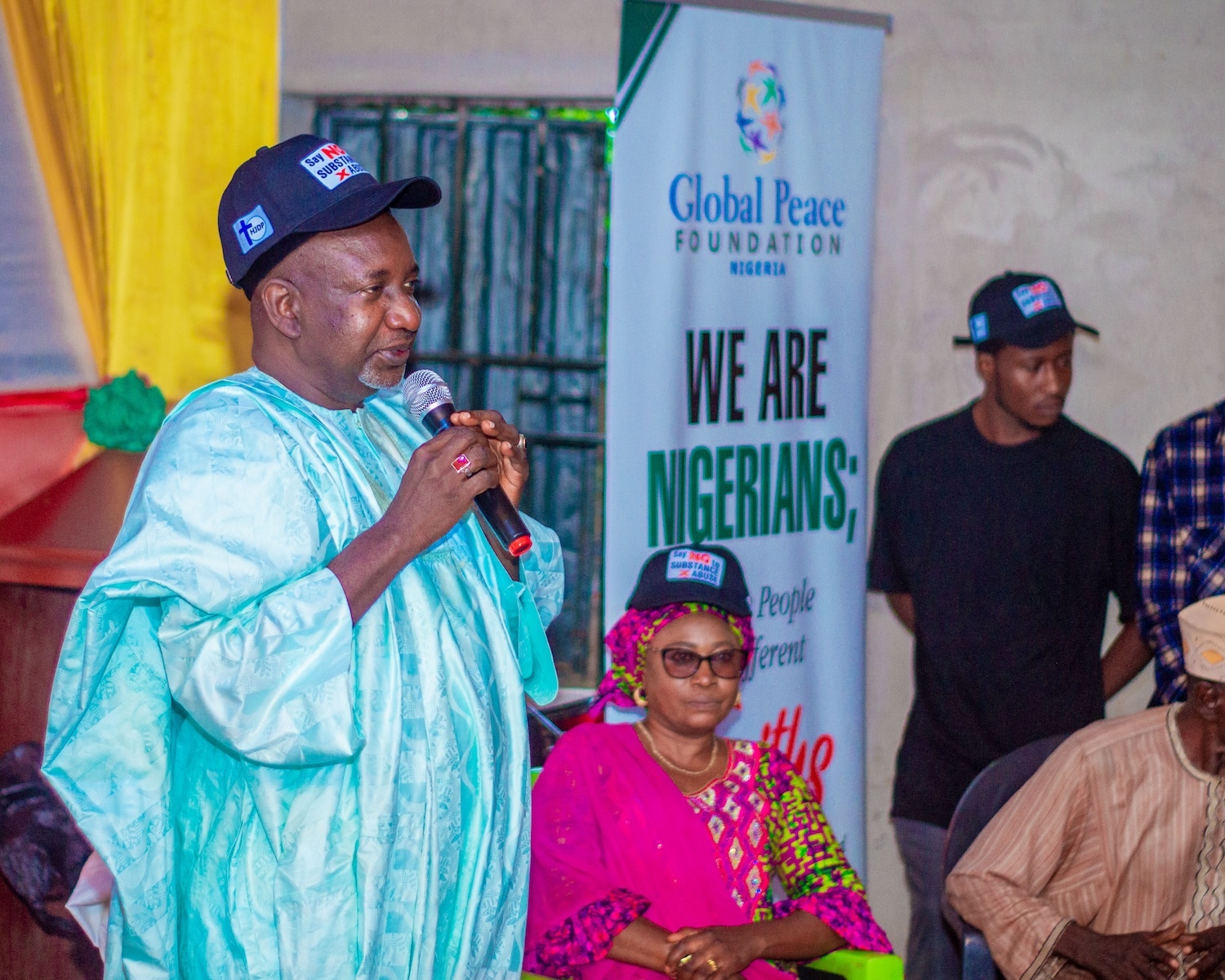 A man in traditional attire speaks into a microphone at an event, empowering youth and promoting peace and resilience, with attendees seated nearby. A banner behind him reads "Global Peace Foundation Nigeria" and "We Are Nigerians".