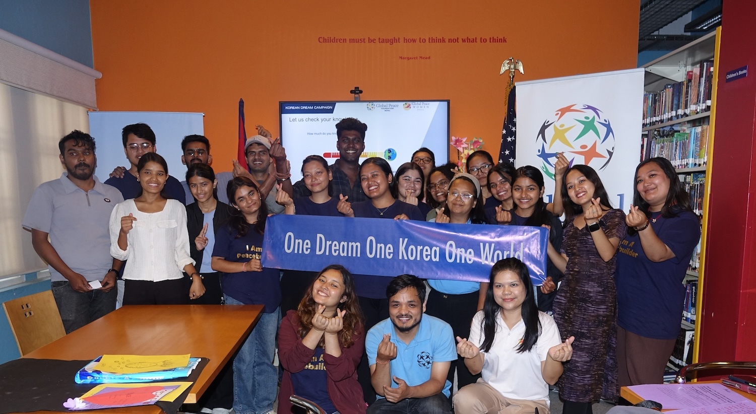 A group of people indoors holding a banner that reads "One Dream One Korea One World." They are smiling and making heart signs with their fingers, celebrating the 2024 International Day of Peace. Library shelves and a sign in the background.