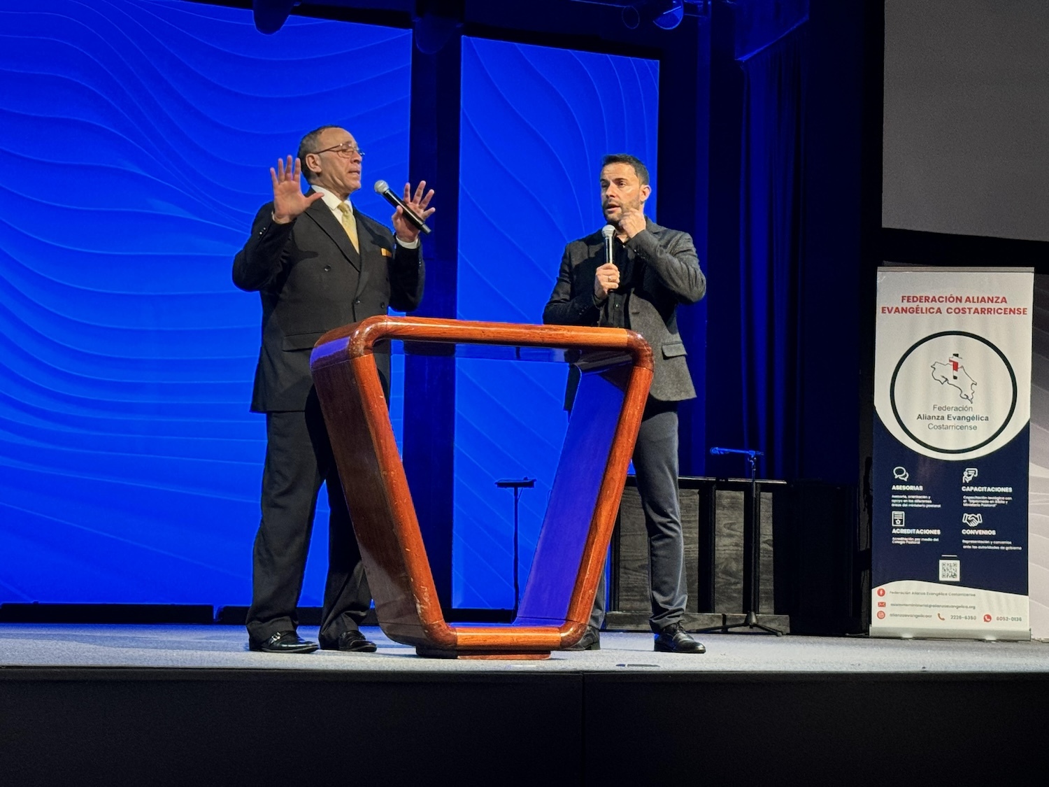 Two men speak on stage behind a wooden podium during an event in Costa Rica. One man, gesturing with both hands raised, passionately discusses religious freedom, while the other holds a microphone. The background features blue wave patterns and a banner.