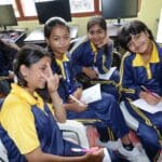 A group of five students in matching uniforms sit indoors, smiling and holding papers and pens, with computers in the background, fostering a culture of peace through character education.