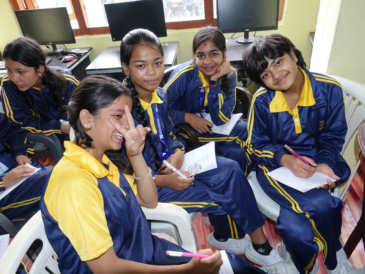 A group of five students in matching uniforms sit indoors, smiling and holding papers and pens, with computers in the background, fostering a culture of peace through character education.