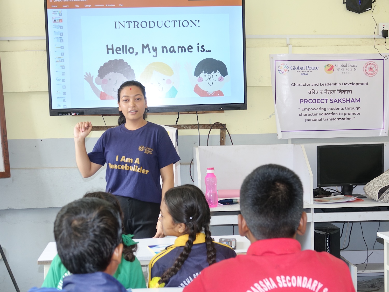 A woman stands in front of a classroom, speaking to a group of students. A projector displays an introductory slide with the text 