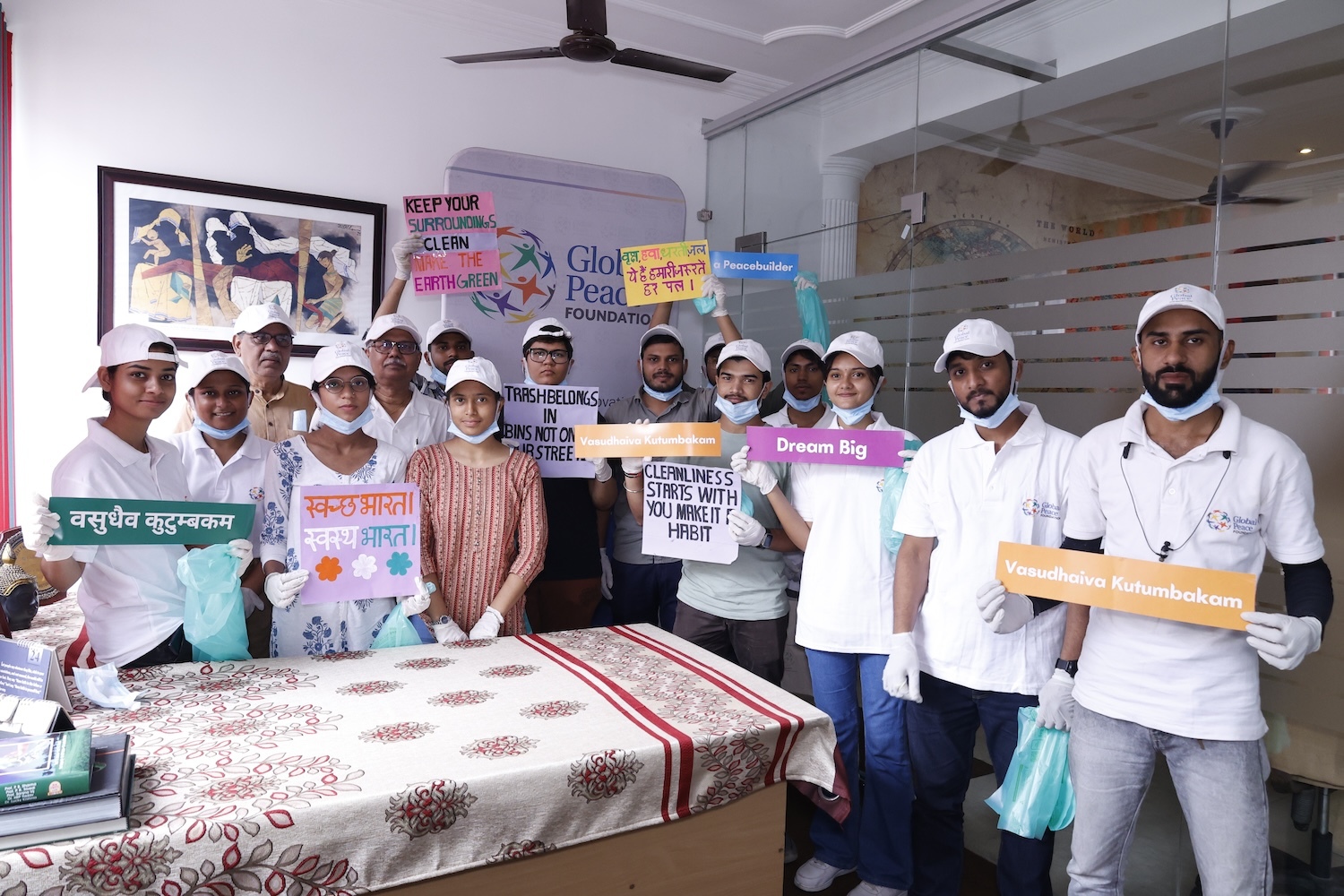 A group of people in white shirts and caps hold signs promoting cleanliness and unity in various languages. They are gathered in a room with a table and posters from the Peace Club advocating for environmental awareness and a Cleaner Future.