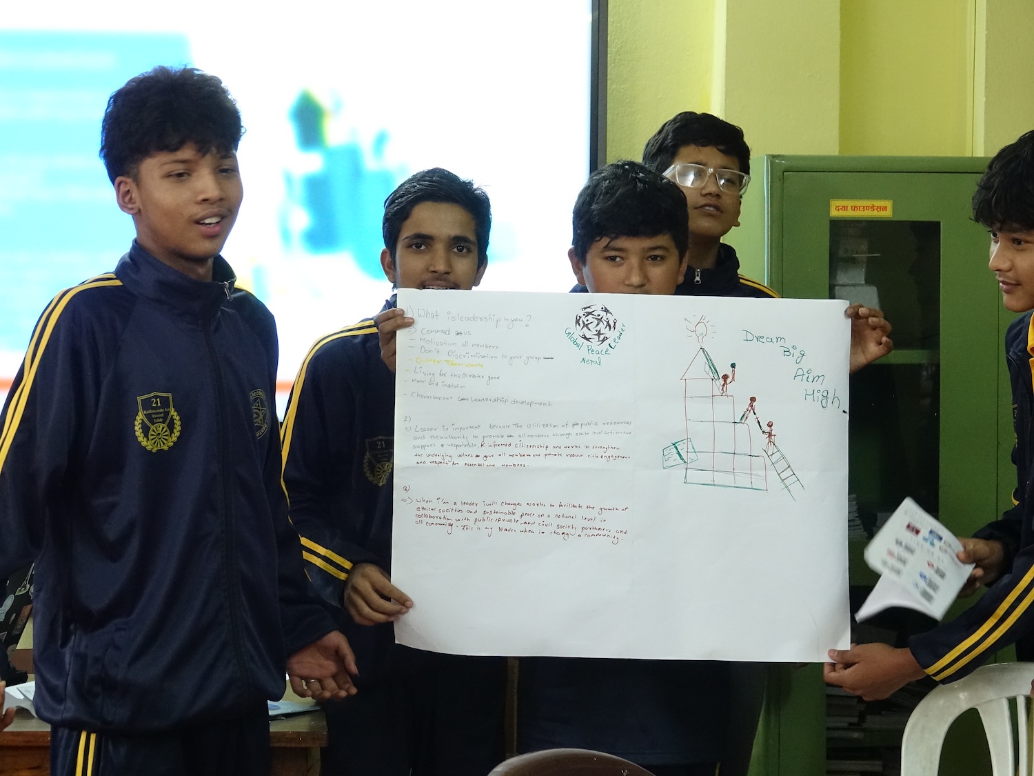 A group of five students in school uniforms presents a handwritten project on a large sheet of paper featuring text and a house drawing, promoting character education through their collective effort.