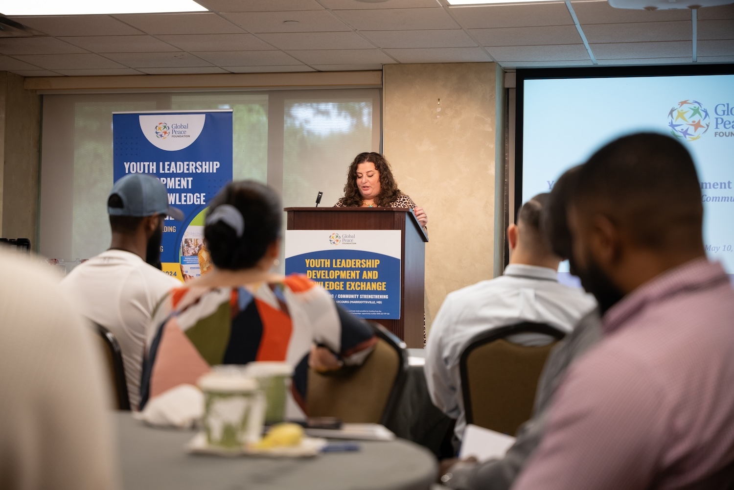 A woman stands at a podium speaking at a conference. The banner reads "Youth Leadership Development and Knowledge Exchange." Attendees are seated, facing her, eager to participate in this vital knowledge exchange.