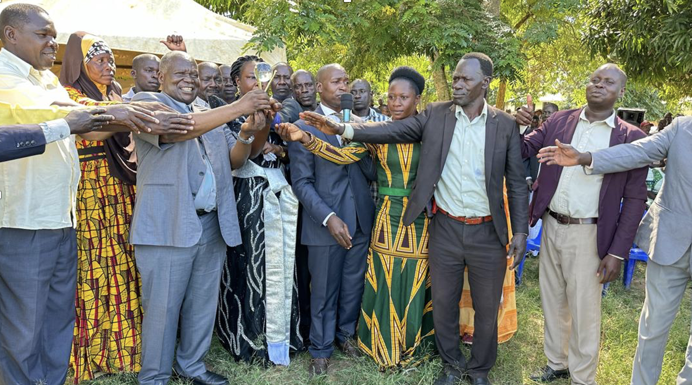 A group of people, some in traditional clothing, stand outdoors in a line, reaching their hands towards the center in a unity or celebratory gesture. The scene reflects the transformative spirit of community and education in Uganda, with lush trees visible in the background.