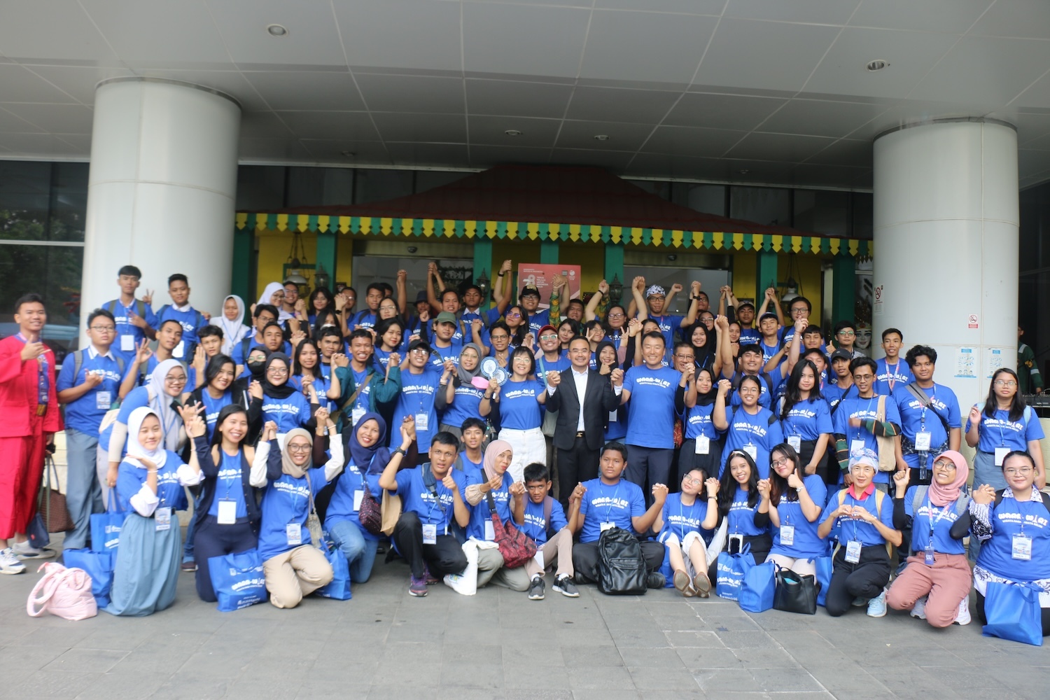 A large group of people, many wearing blue shirts and holding blue bags, pose together in front of a building in Indonesia, smiling and giving thumbs up.