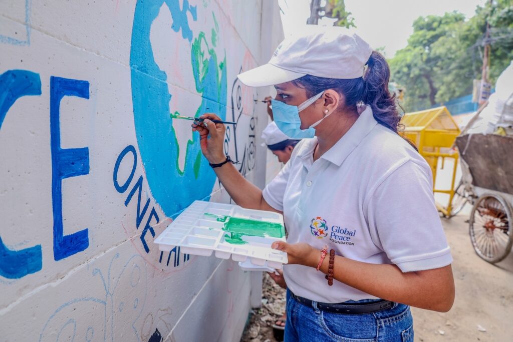 A woman wearing a mask and a cap paints a mural of the Earth on a wall. She holds a paint palette with green paint and works on the continents with a brush, representing the mission of the Peace Club for a Cleaner Future. Another person is painting in the background.