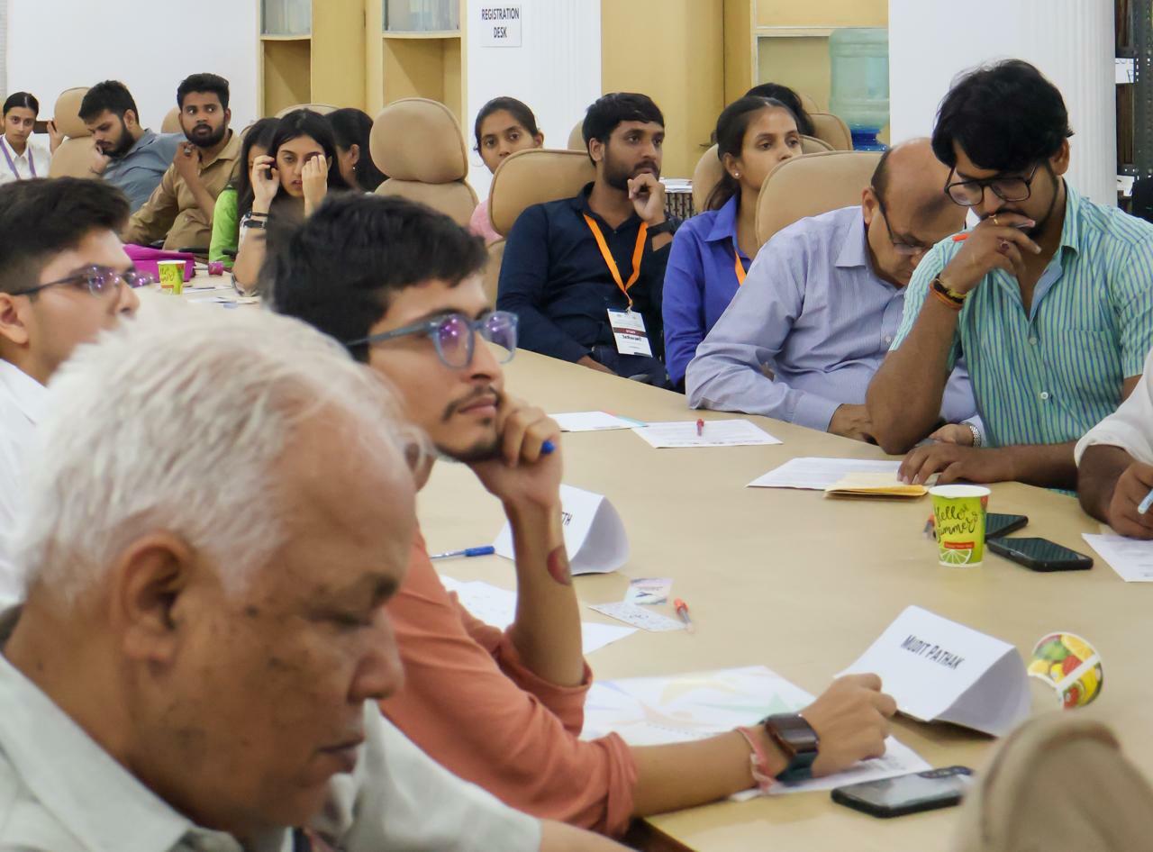 People seated around a table at a meeting, attentively listening and taking notes. Various papers, cups, and office supplies are visible on the table as they discuss sustainability initiatives in honor of International Youth Day.