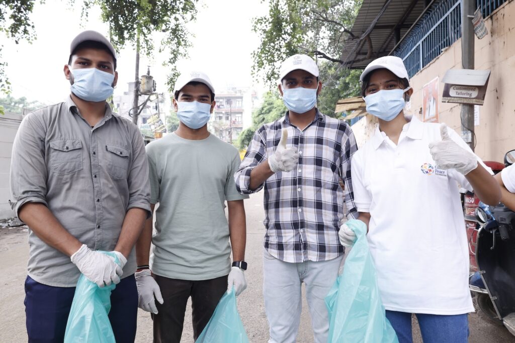 Four people standing outdoors, wearing masks, gloves, and holding trash bags; they pose for the camera with thumbs-up gestures, indicating their participation in a Cleaner Future community clean-up event.