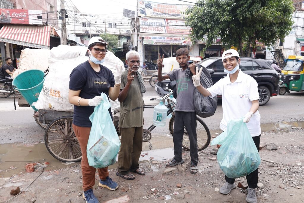 A group of people from the Peace Club, wearing gloves and holding trash bags, stand on a street with a rickshaw and a bicycle in the background, engaging in a cleanup activity for a cleaner future.