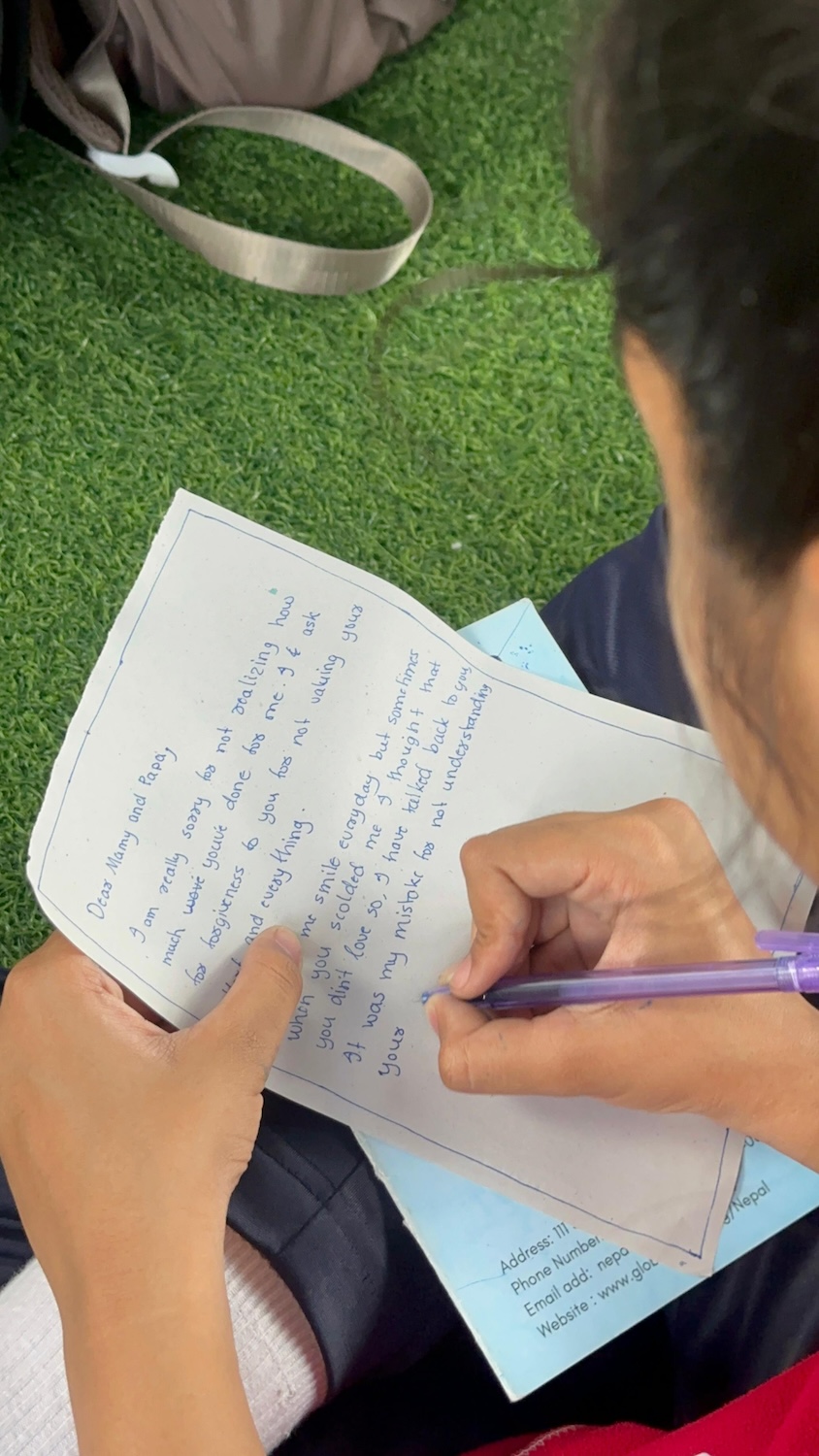 A person is writing on a piece of paper with a purple pen while seated on green grass, promoting a Culture of Peace. Their wrist rests on a blue folder beneath the paper, and a brown backpack is visible in the background.