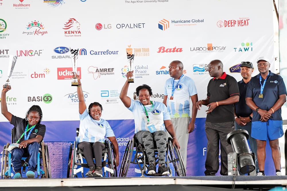 A group of wheelchair athletes proudly stands on stage, two holding trophies, with several sponsors' logos in the background. The event celebrates their achievements and highlights partnerships, including GPF Kenya and the Standard Chartered Nairobi Marathon.