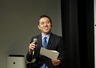 A person in a suit holds a microphone and a paper, smiling warmly while seated indoors, as they prepare to discuss the International Day of Peace 2024 event in Japan.