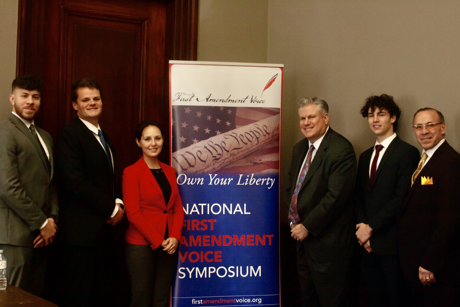 Six people stand next to a banner that reads "National First Amendment Voice Symposium: Own Your Liberty," with a partial image of the U.S. Constitution, celebrating free speech.