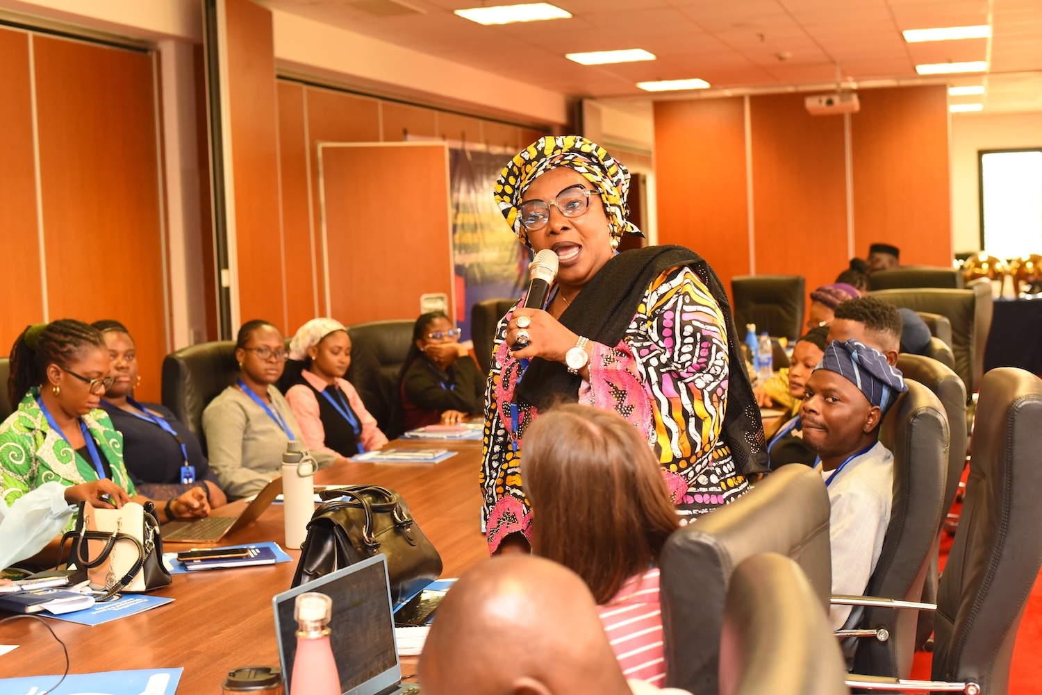 At a conference table in Nigeria, a woman speaks into a microphone, leading an interfaith dialogue. Surrounded by attendees seated and listening attentively, she champions the cause of religious tolerance with eloquence and wisdom.