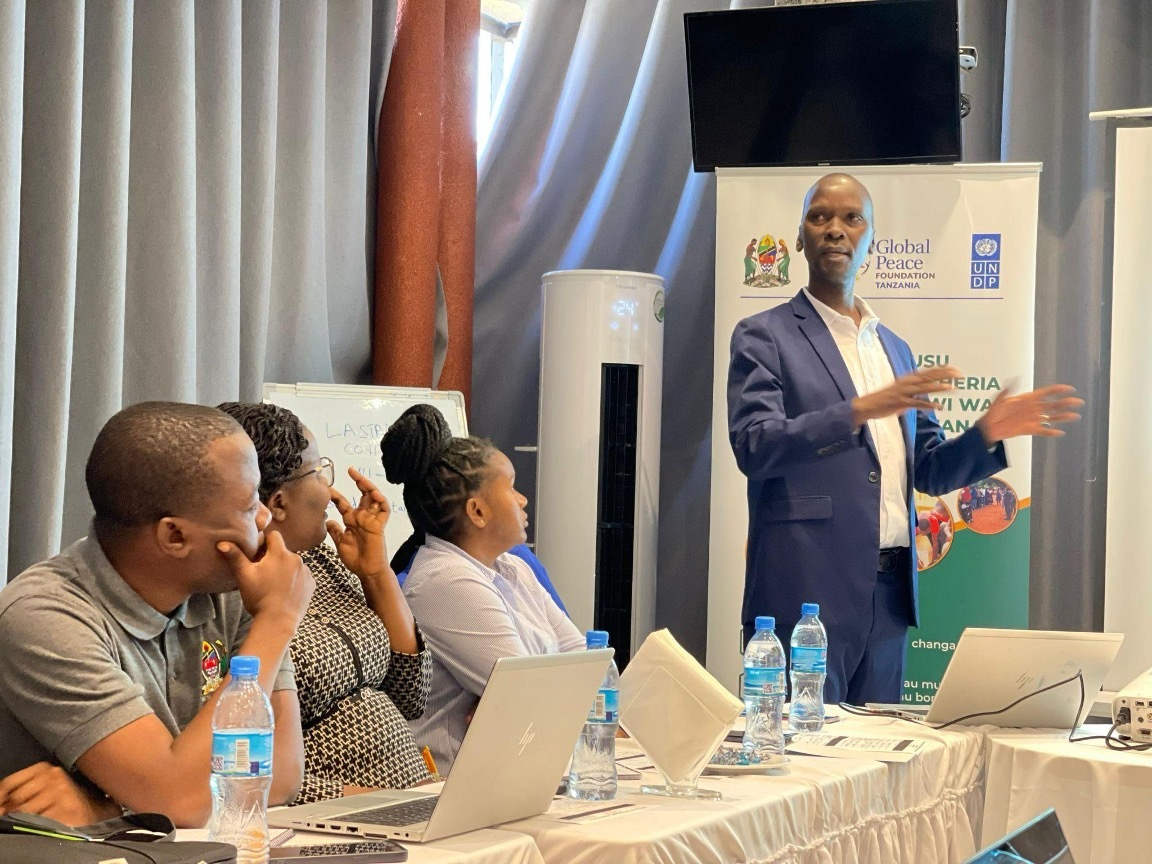 A man stands speaking to a seated audience in a conference room, passionately addressing participants of the Young Leaders and Peace and Youth Program. Laptops, water bottles, and a projector screen are visible as the session unfolds, fostering new ideas for youth empowerment projects in Tanzania.