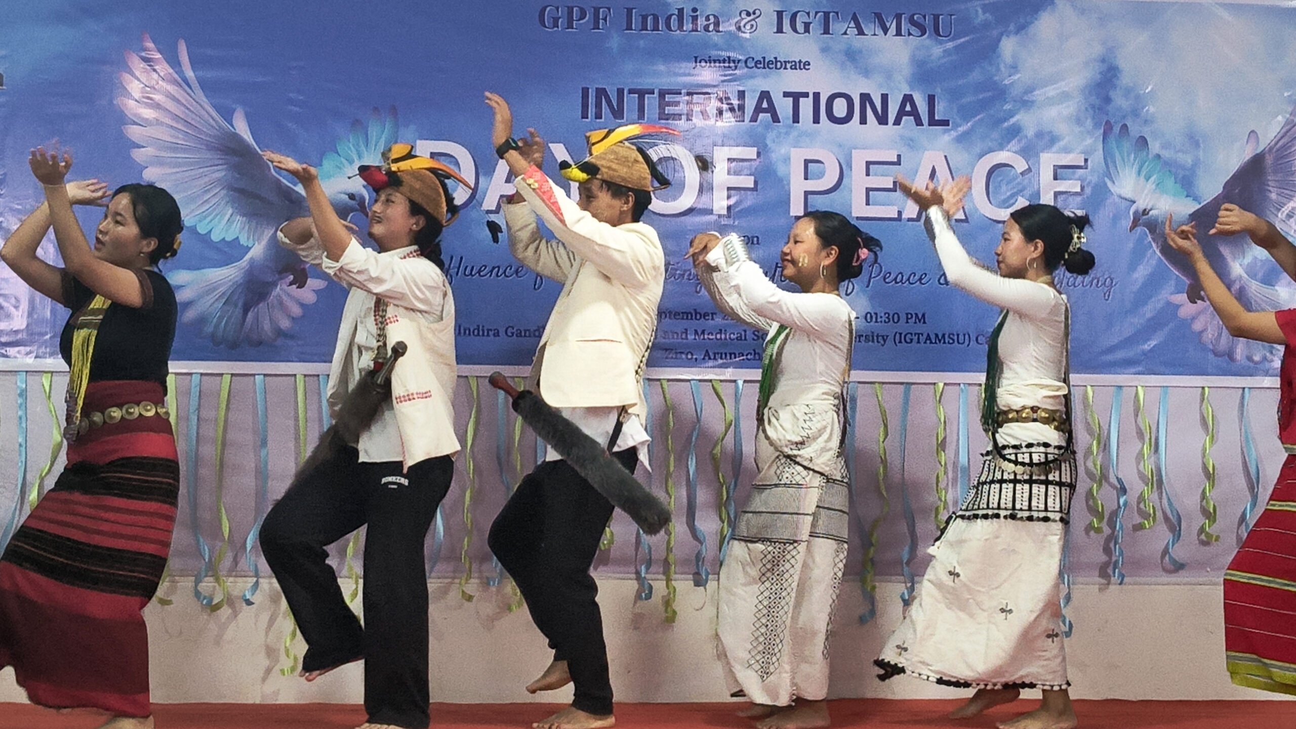 A group of people in traditional attire performs a cultural dance on stage, capturing the confluence of cultures, with a banner reading "International Day of Peace" in the background.