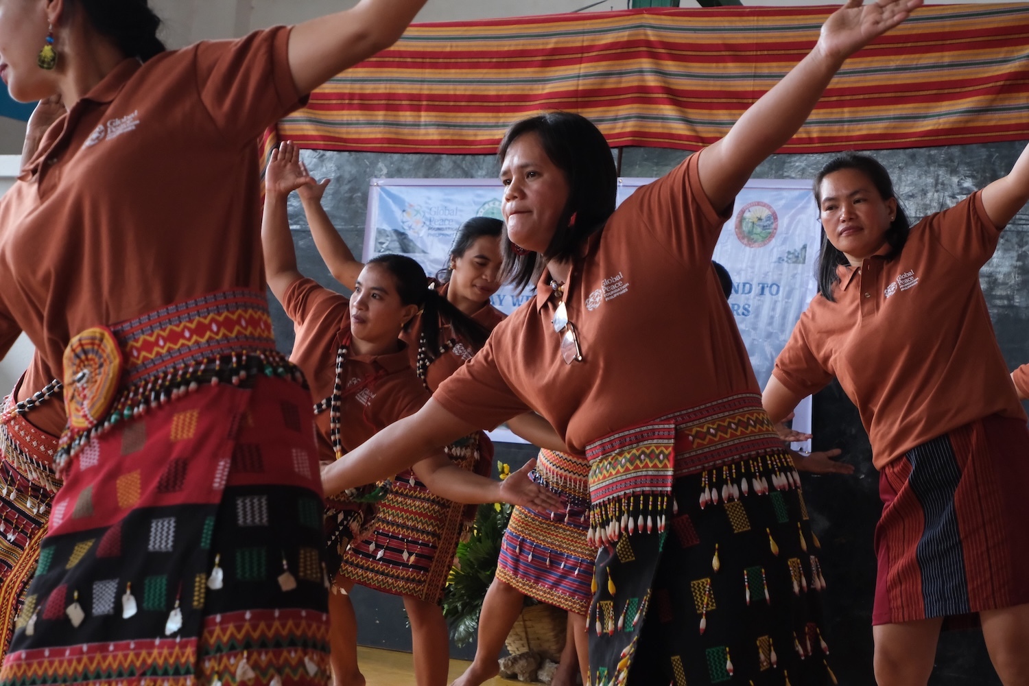 A group of people wearing traditional clothing perform a dance in a room adorned with banners, celebrating the Chumma-ay Weavers and Bead-makers Association.