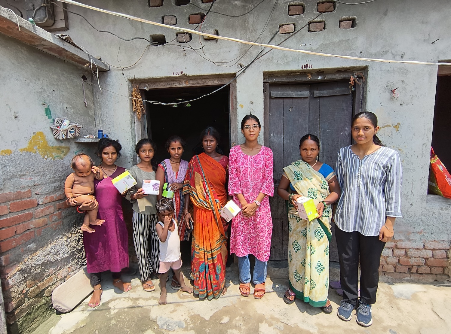 Seven women and a child stand outside a building, some holding books, in an informal and sunny scene. With brick and concrete walls hinting at the urban slum setting, they're likely participants of a GPF India initiative focused on menstrual health awareness.