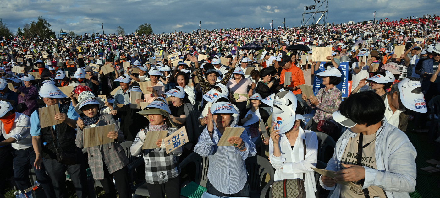 A large outdoor crowd, mostly wearing hats, holds wooden plaques in the air at the International Forum. The sky is partly cloudy.