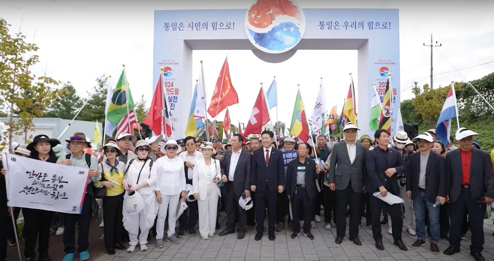 A diverse group of people holding various national flags stand together under an archway with banners, symbolizing unity at the International Forum on One Korea. Among them, the spirit of a Unified Korea shines brightly, echoing the aspirations of the Korean Dream Grand March.