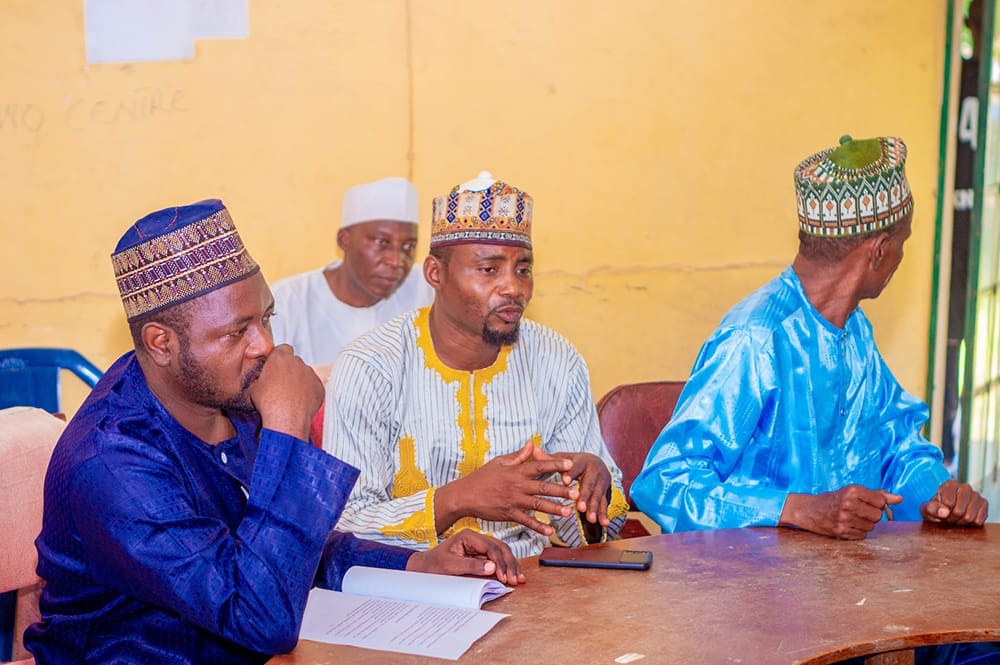 Four men in traditional attire sit at a table in discussion. A man in a blue outfit takes notes while another in a striped shirt gestures passionately about equitable access to education for vulnerable girls. A yellow wall forms the backdrop, reflecting the vibrant spirit of Kaduna State.