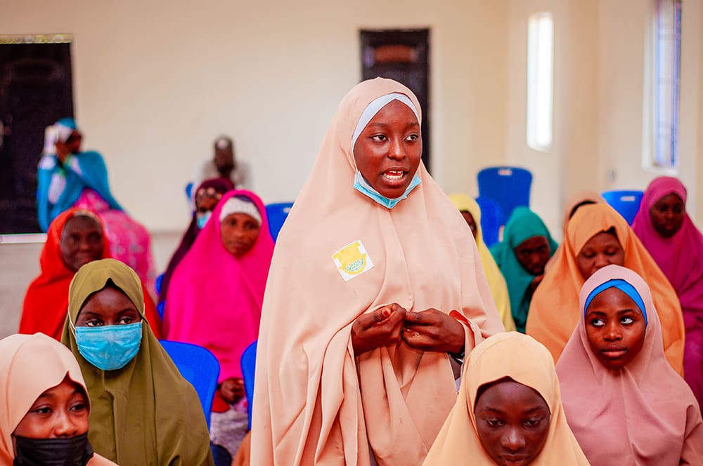 In Kaduna State, a group of girls in colorful hijabs gather indoors. One stands to speak, representing their drive for equitable access to education. Blue chairs form the backdrop as they discuss their aspirations and challenges.