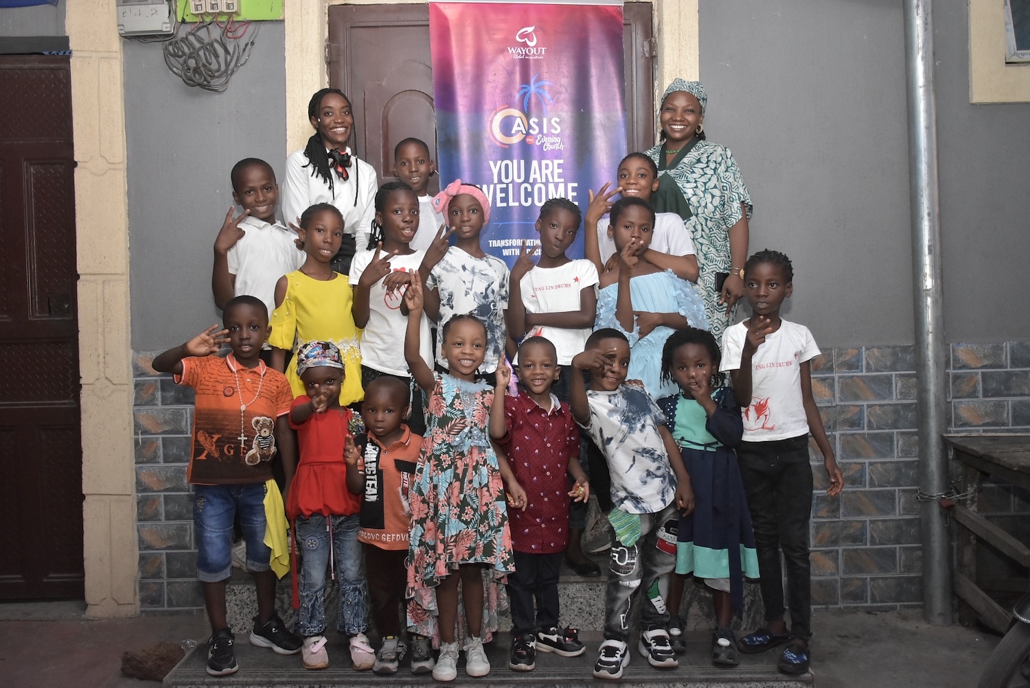 A group of children and two adults stand before a "You Are Welcome" banner, capturing the spirit of the International Day of Peace. Smiling for the photo, they embody unity and warmth.