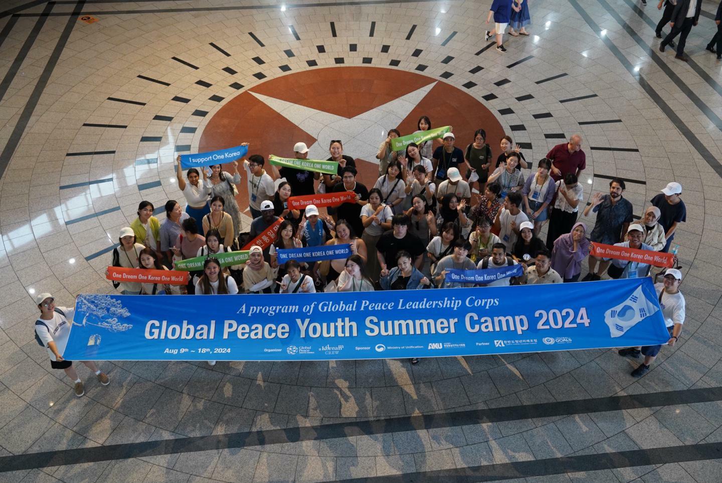 A group of people stands on a patterned floor indoors, holding colorful banners and a large blue one reading "Global Peace Youth Summer Camp 2024," celebrating leadership and youth empowerment through cultural exchange.