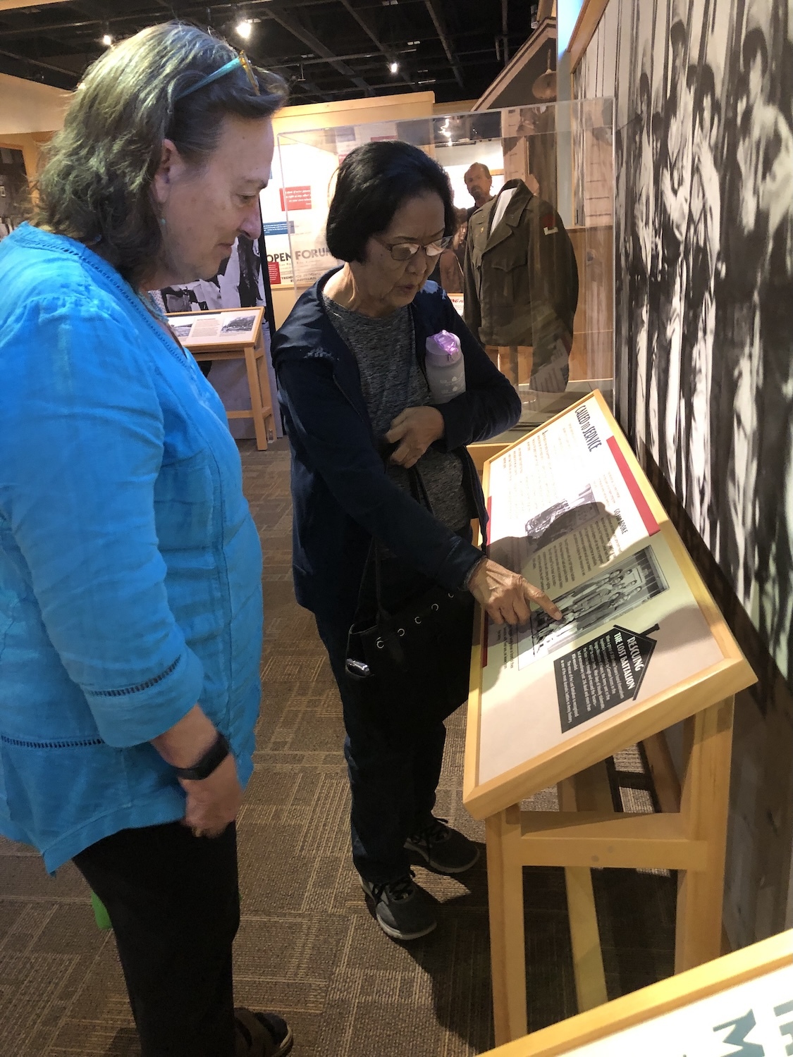 Two people examine an exhibit display in a museum, exploring historical photographs and text on a wooden stand that capture the essence of the Heart Mountain Experience.