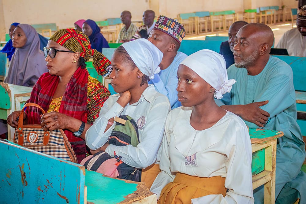 In a classroom setting, a group of people seated in rows of wooden benches attentively face forward, embodying the pursuit of education and equitable access for all, including vulnerable girls.