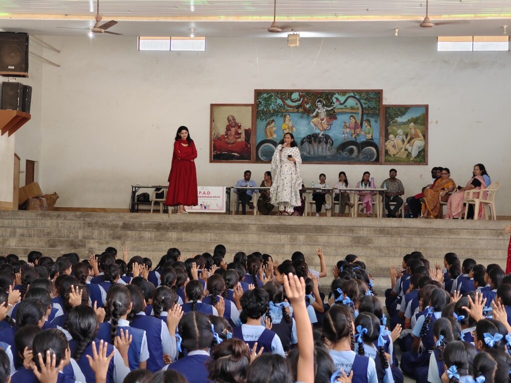 Two women stand on stage, educating a group of students in uniforms about menstrual health. The engaged students have their hands raised as paintings hang on the wall behind them, showcasing the impactful event organized by GPF India.