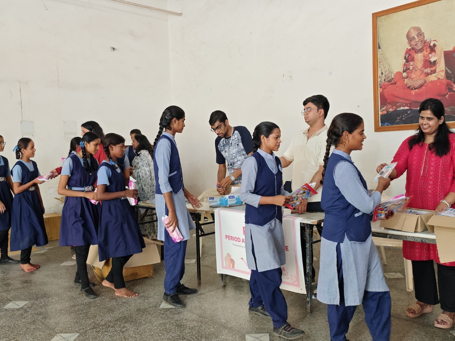 Students in uniform receive supplies focused on menstrual health from tables staffed by adults in a room with a large, framed portrait on the wall, as part of GPF India's initiative to educate and support their well-being.