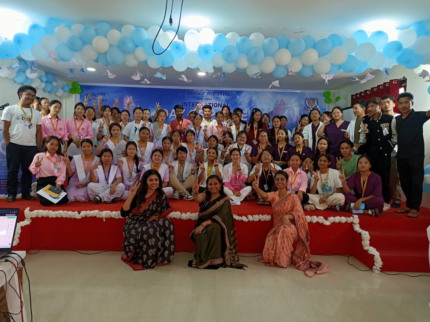 A group of people in martial arts uniforms and traditional attire poses on stage with medals and certificates, surrounded by blue and white balloon decorations. Their unity reflects a commitment to peacebuilding through leadership training.