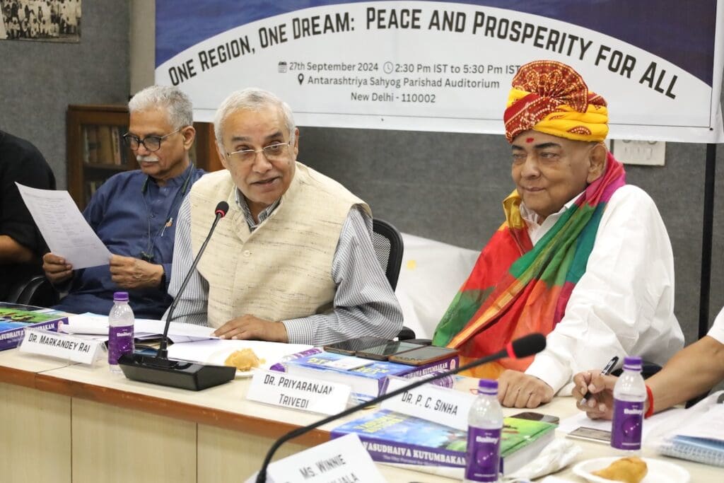 A panel of three men sits at a conference table with microphones in front of them. The banner behind reads