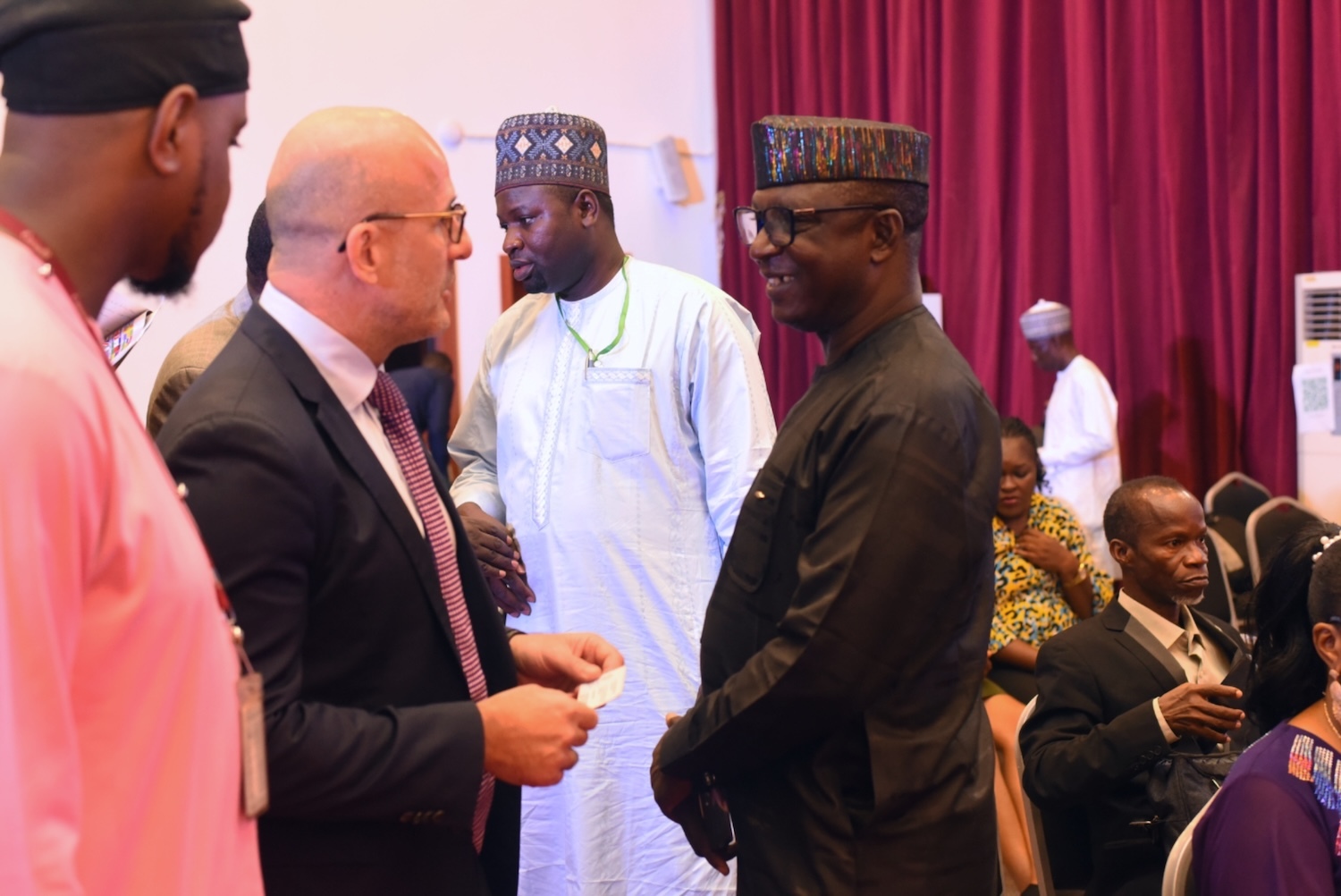 A group of people in formal and traditional attire engage in conversation about peacebuilding in Nigeria, set against a room with red curtains.