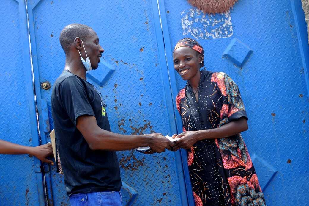 Two people stand in front of a blue gate in Nigeria. One is handing something to the other, who is smiling warmly, showcasing the spirit of cooperation fostered by Makeover Initiative.