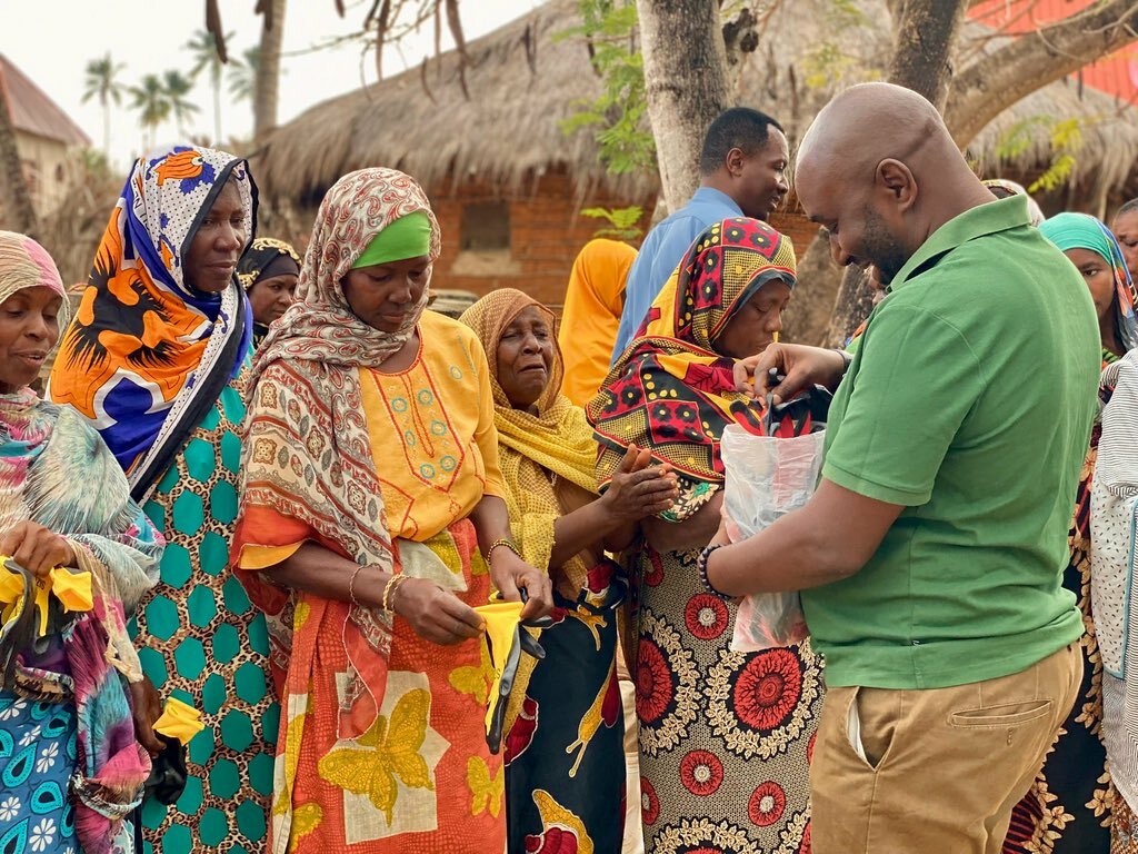 A man in a green shirt, as part of an Environmental Outreach initiative, distributes items to a group of Tanzanian women in colorful attire standing in line outdoors.