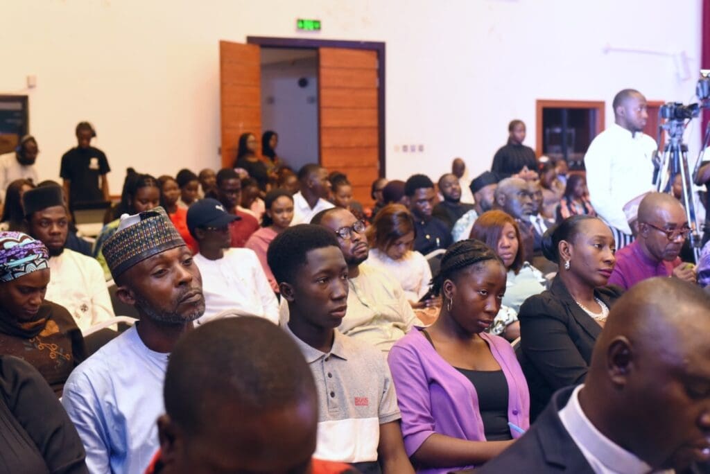 A diverse audience seated in a conference room, listening attentively to insights on peacebuilding. Some people are taking notes, as a camera captures the moment on the right side, highlighting efforts in Nigeria.