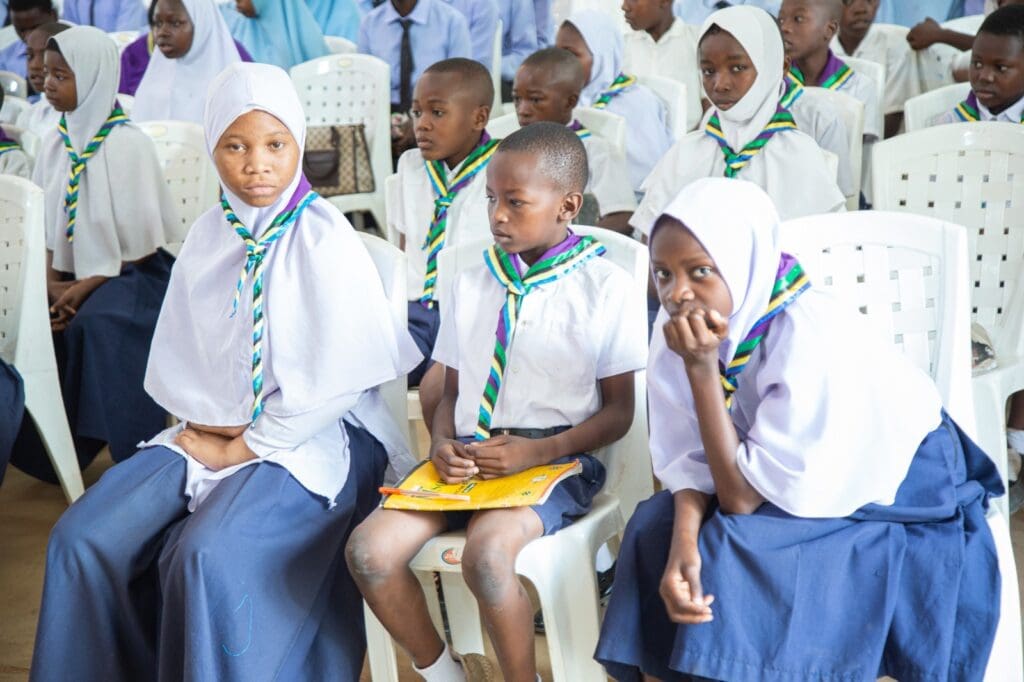 A group of Tanzanian students in uniforms and headscarves, wearing scout neckerchiefs, are seated on white chairs. Their gathering reflects a spirit of unity reminiscent of the International Day of Peace.