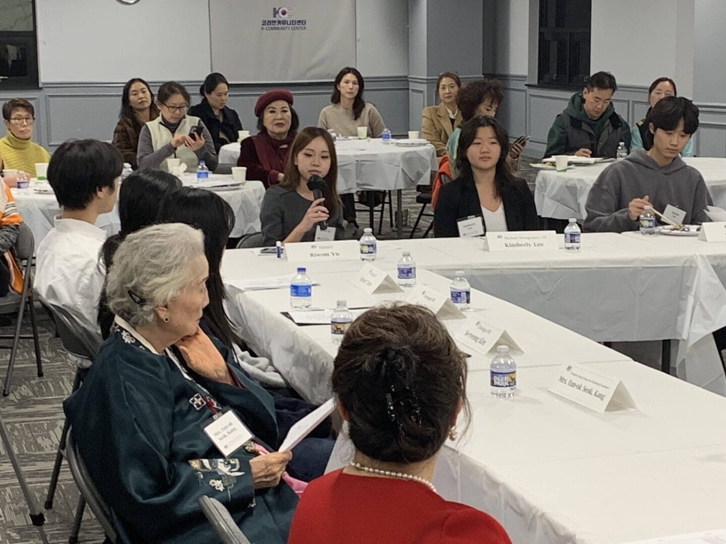 In a meeting room filled with people seated at tables, some displaying AKU-Washington name tags, a woman holds the microphone, engaging them in a dynamic Talk Contest. Bottles of water and papers scatter across the tables as attendees listen attentively.