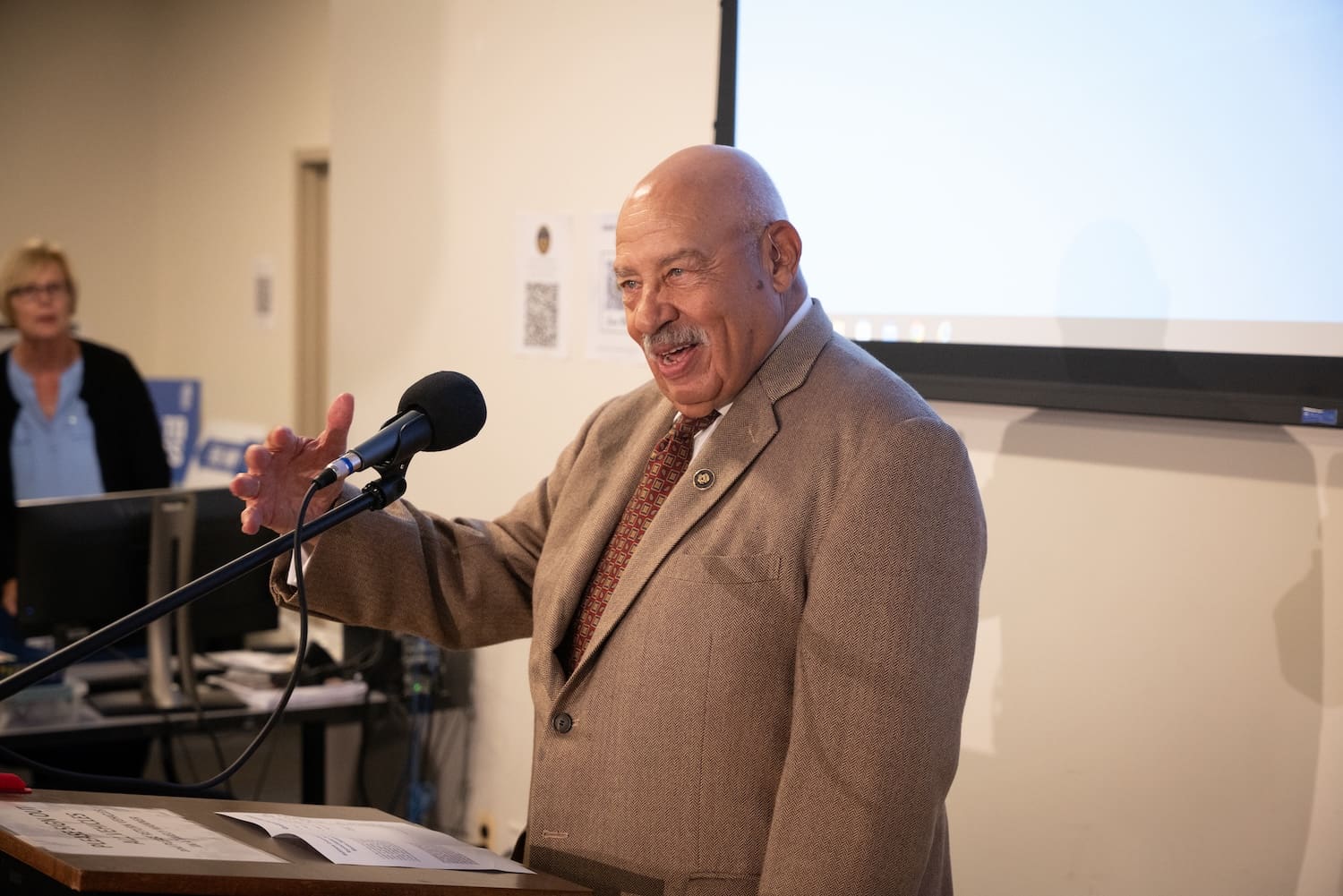 A man in a suit speaks at a podium with a microphone, addressing Baltimore residents about race relations, as a woman stands in the background in front of a projected screen.
